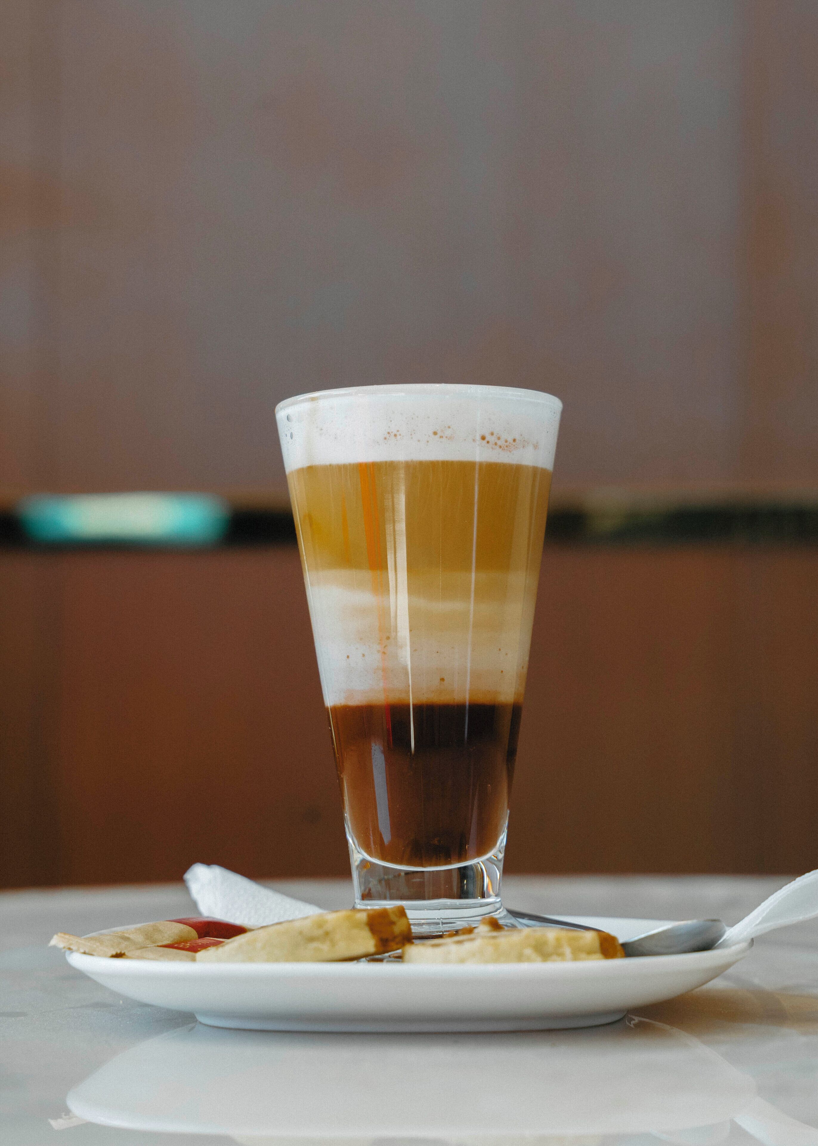 Un verre contenant une boisson à plusieurs couches, posé sur une assiette blanche avec une cuillère, le tout placé sur une table devant un arrière-plan flou.