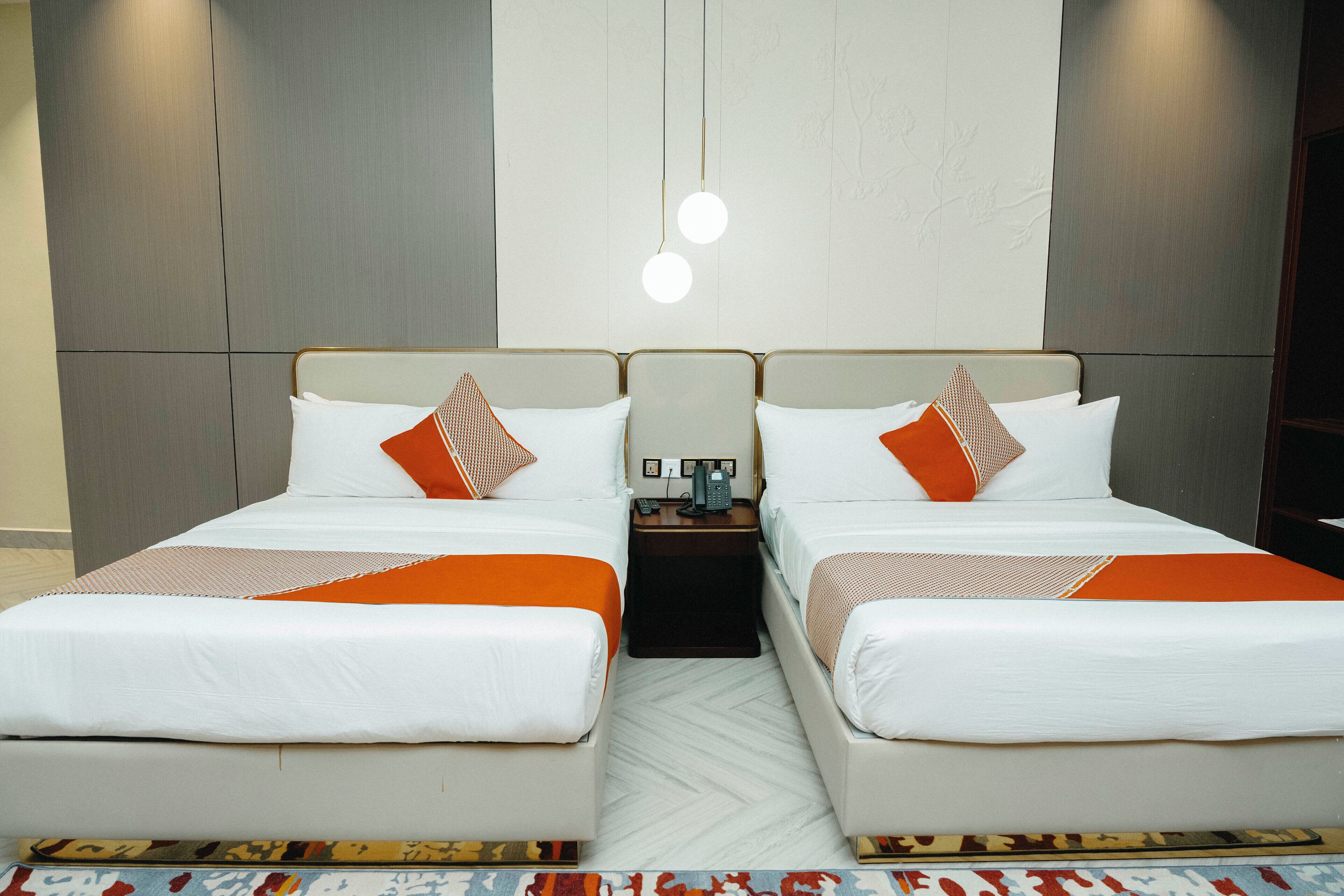 Habitación doble con dos camas, cada una con ropa de cama blanca y detalles en naranja, una mesita de noche pequeña con teléfono y una lámpara de pared.