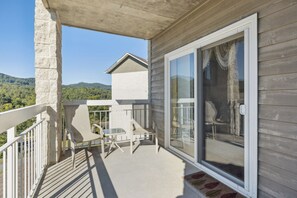 Un balcon donnant sur un paysage de montagnes boisées, avec une balustrade blanche et une porte-fenêtre coulissante.