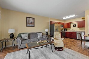 A living room with a sofa, coffee table, and a chair. There is a kitchen area with cabinets and appliances.