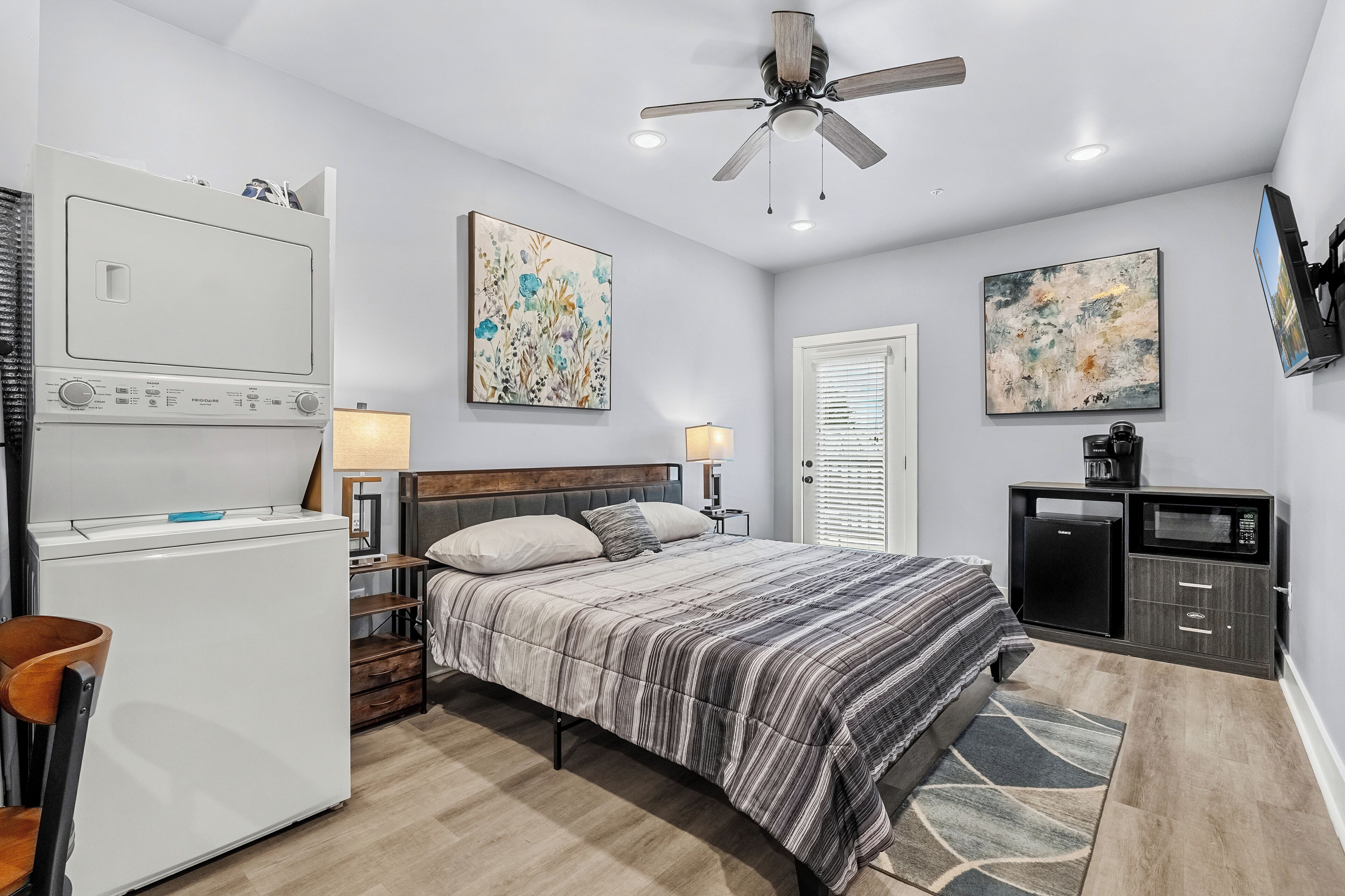 A hotel room with a bed, a desk, a refrigerator, a ceiling fan, and artwork on the walls.