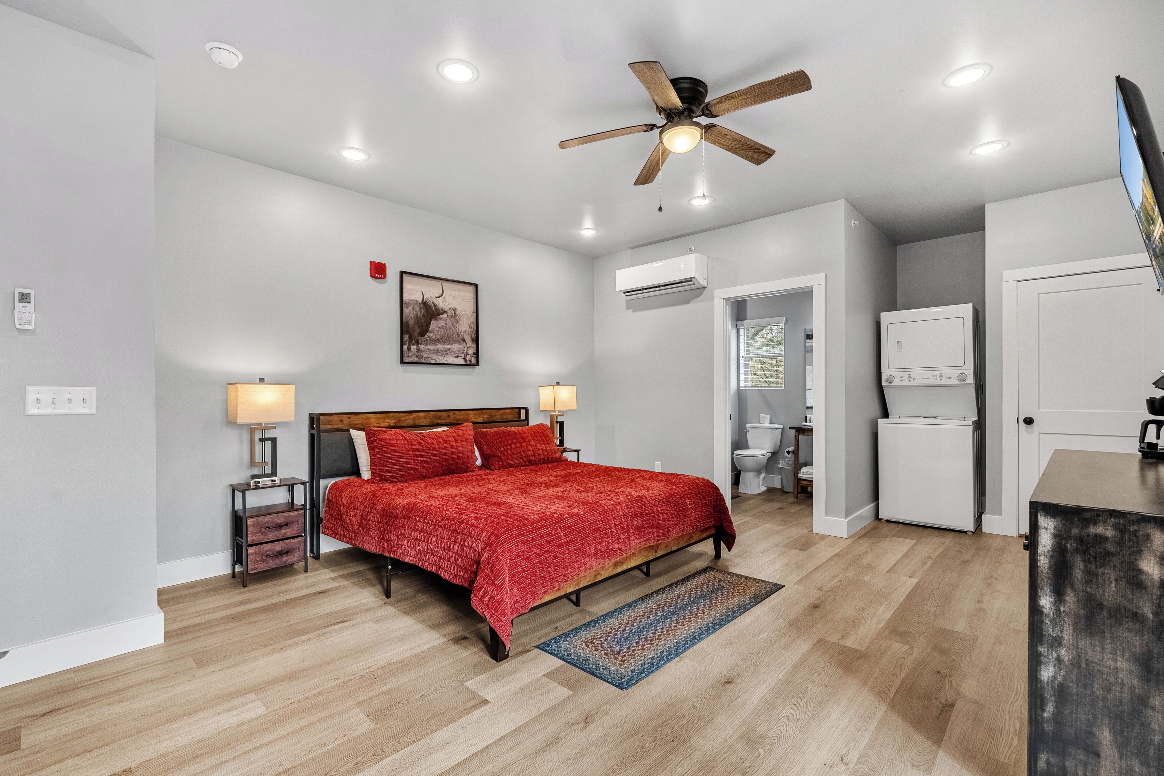 A bedroom with a bed, bedside lamps, a ceiling fan, an air conditioning unit, and a door leading to a bathroom.