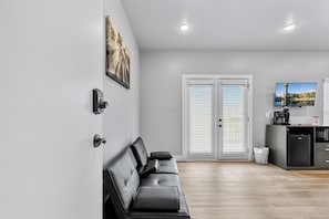 A modern living room with a black leather sofa, a painting, a door with white shutters, and a kitchen area with a refrigerator and a coffee machine.