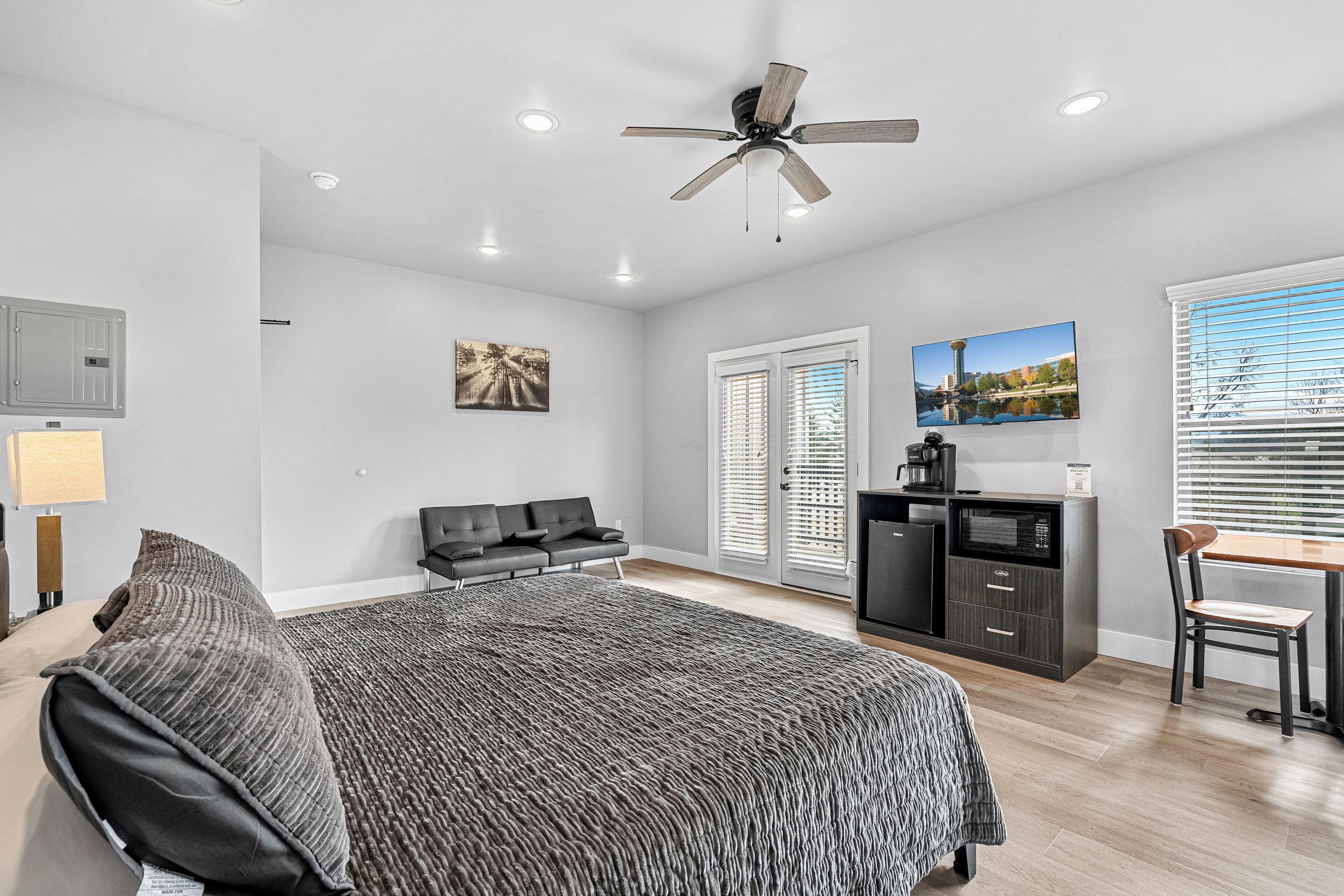 A modern bedroom with a bed, a desk, a chair, a ceiling fan, and a large window with blinds.