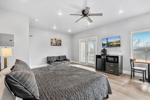 Een moderne slaapkamer met een bed, een bureau, een stoel, een plafondventilator en een groot raam met jaloezieën.