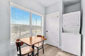 Een kleine keuken met een eettafel en stoelen, een witte koelkast, en een raam met jaloezieën.