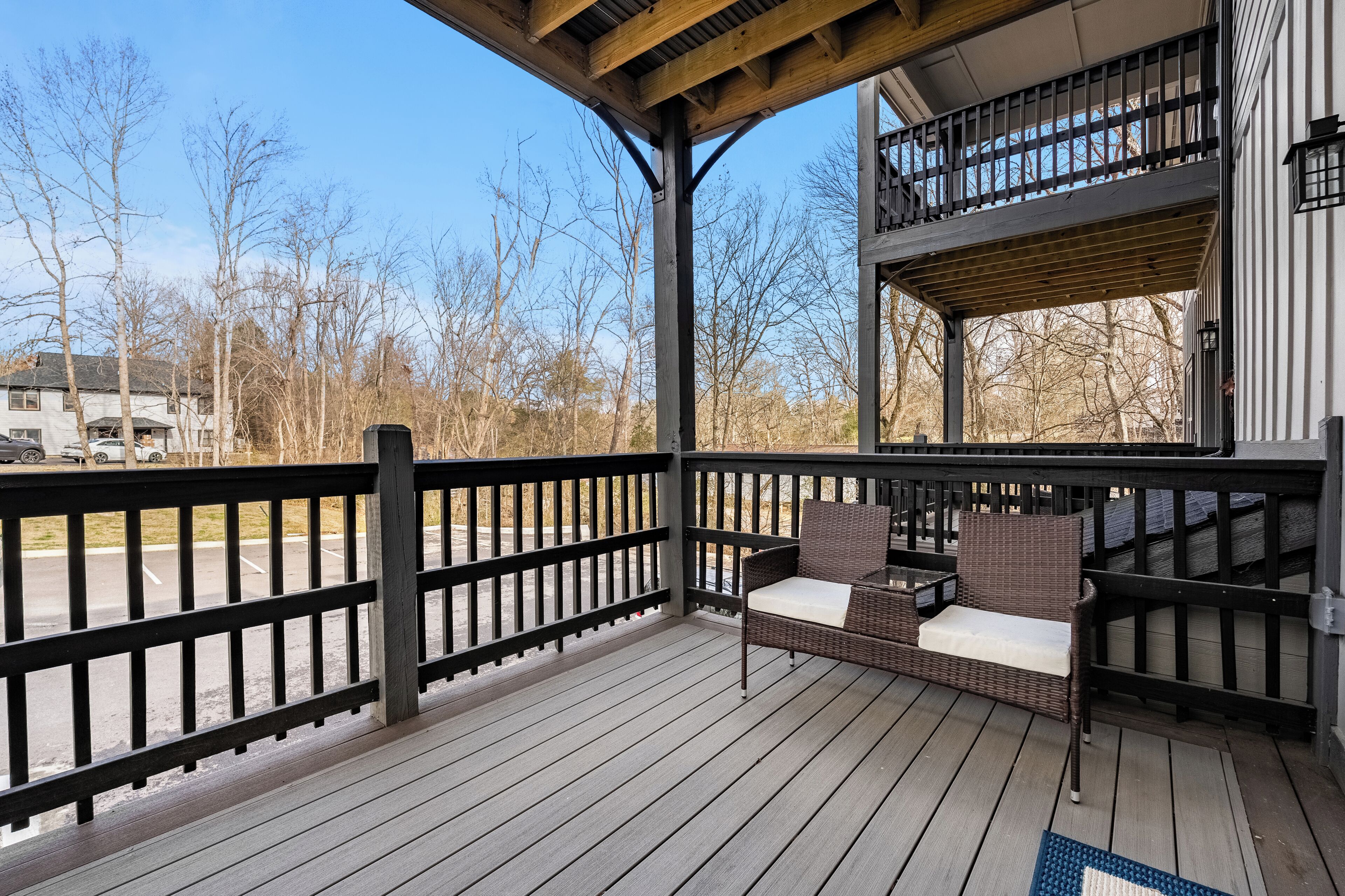 A wooden deck with a sofa and a table, overlooking a parking area and trees.