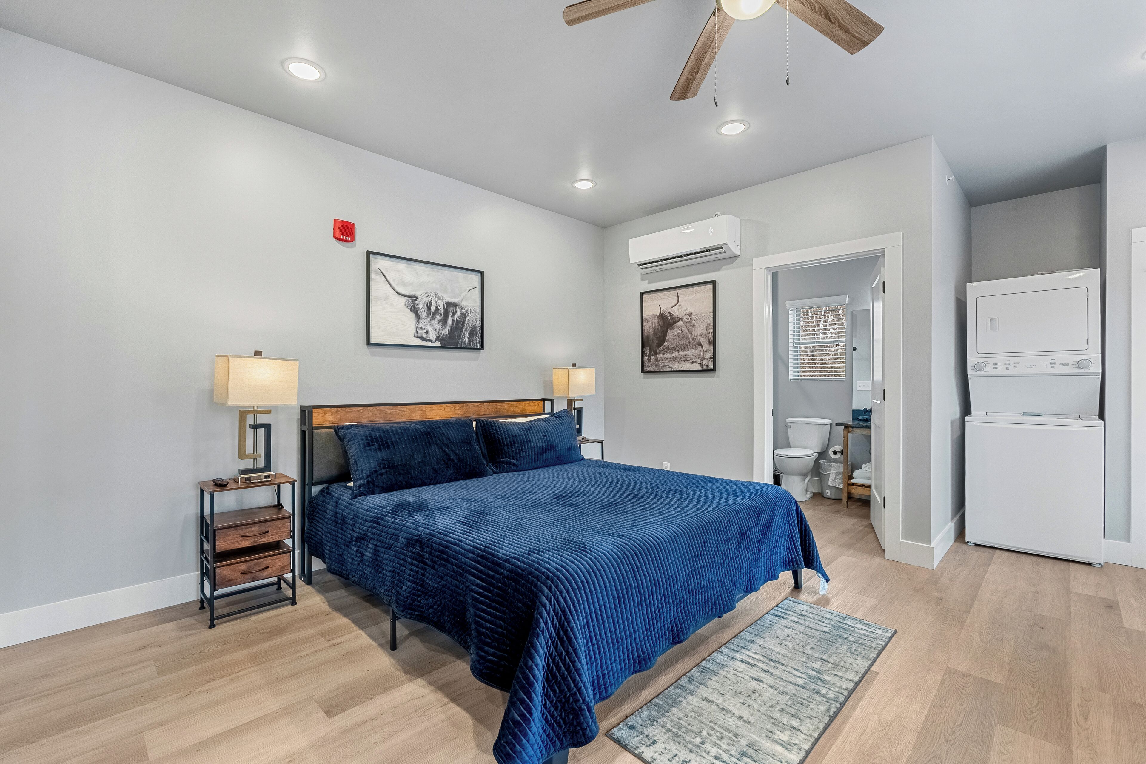 A bedroom with a bed, bedside tables, a ceiling fan, wall-mounted air conditioning, and a door leading to a bathroom.