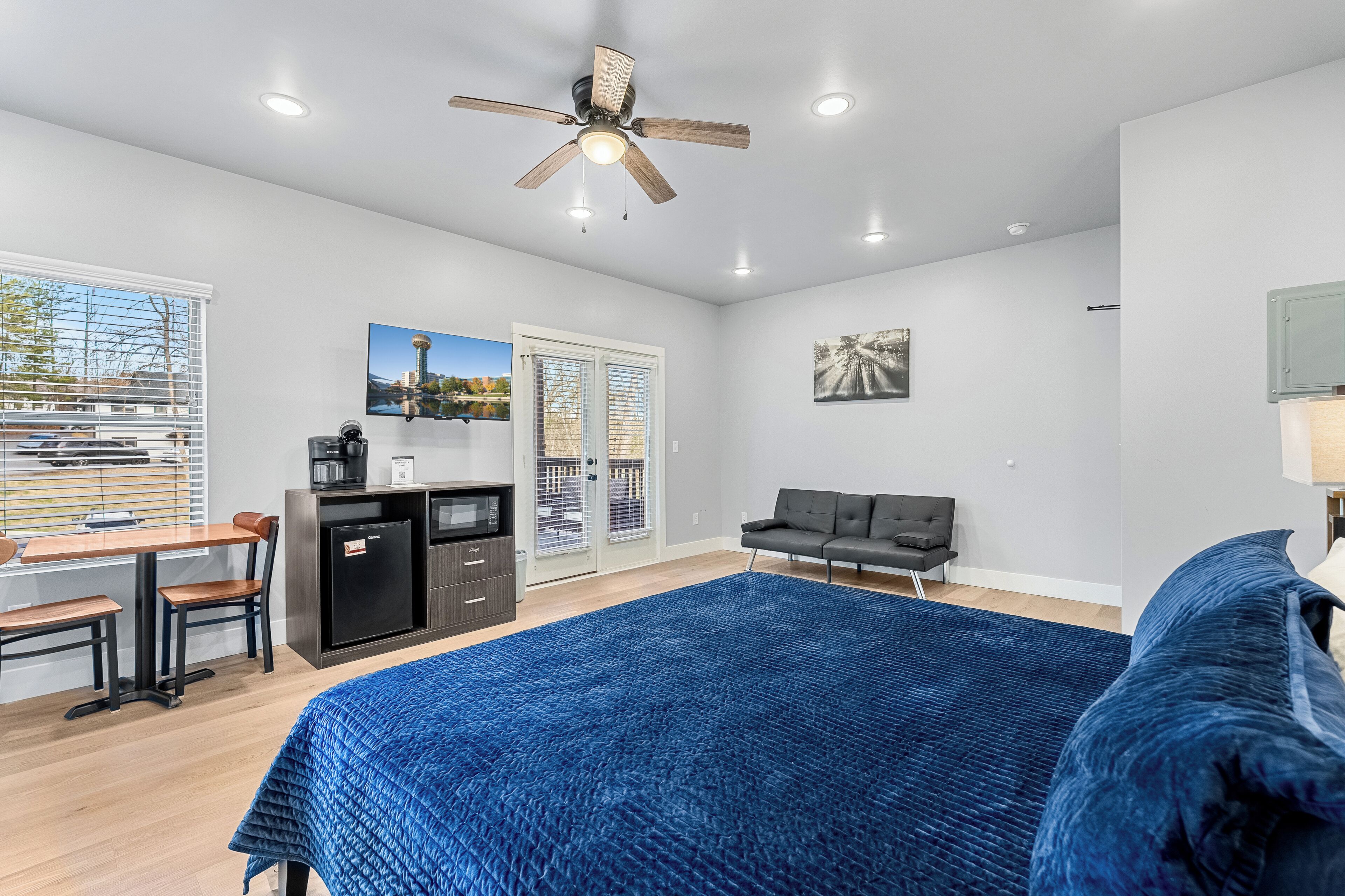 A modern bedroom with a large bed, a desk with a chair, a sofa, a ceiling fan, and a view through the window.