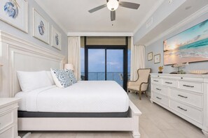 A bedroom with a bed, a dresser, a chair, a ceiling fan, and a large window with a view.