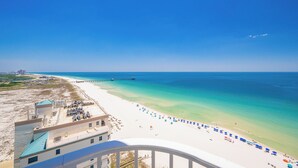 Blick auf einen Strand mit einer Steg, der sich ins Wasser erstreckt, feinem Sand, Sonnenschirmen und einem Gebäude mit blauem Dach.