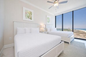Un dormitorio con dos camas, ventilador de techo, obras de arte en las paredes y un ventanal con vista a la playa.