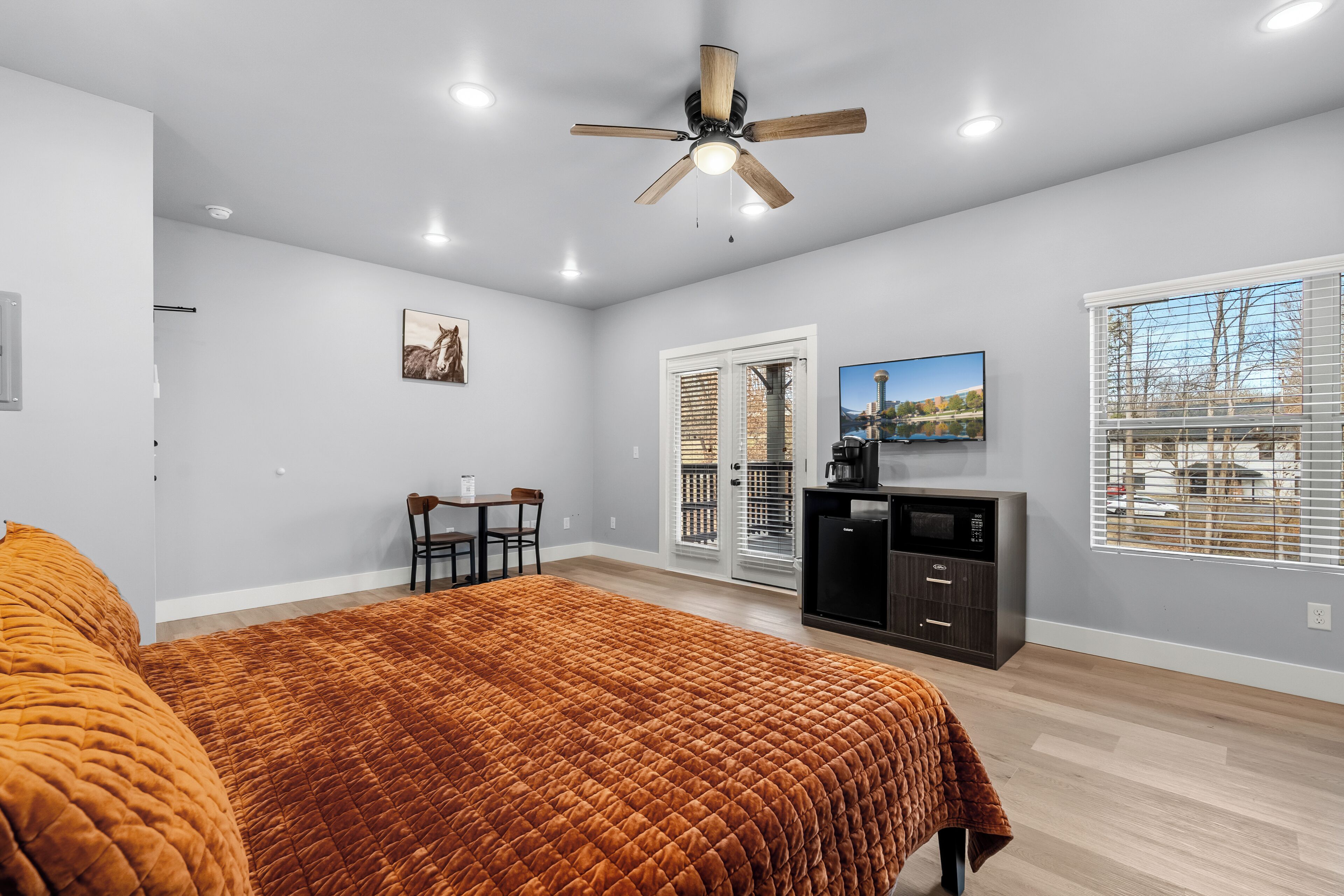 A bedroom with a bed, a ceiling fan, a desk with chairs, a television, and a view of the outside through a window.