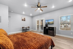 A bedroom with a bed, a ceiling fan, a desk with chairs, a television, and a view of the outside through a window.