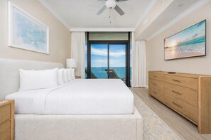 A hotel room with a large bed, a wooden dresser, a flat-screen TV, and a balcony with a view of the ocean.