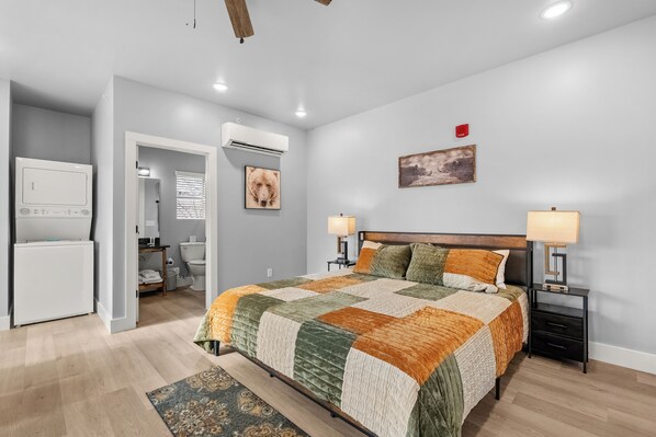 A bedroom with a bed, bedside lamps, a nightstand, a wall-mounted air conditioner, a framed picture, and a small table with a lamp.