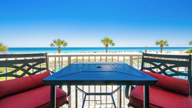 Un balcon avec une table et des chaises, donnant sur une plage bordée de palmiers et sur l’océan.