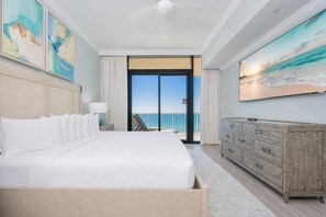 A hotel room with a large bed, a wooden dresser, and a balcony with a view of the ocean.