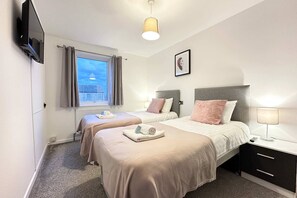 Une chambre d’hôtel avec deux lits, une télévision, une fenêtre avec des rideaux, un tableau au mur et une table de chevet avec une lampe.