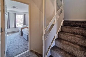 Une chambre à coucher avec un lit, une fenêtre avec des rideaux, un sol recouvert d’une moquette et un escalier muni d’une main courante.