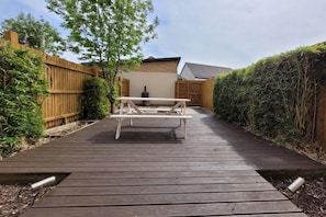 Eine Holzterrasse führt zu einem weißen Picknicktisch mit einem Grill im Hintergrund.