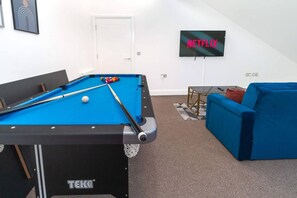 A pool table, a blue sofa, a television displaying 'NETFLIX,' and a door in a room with carpeted flooring.