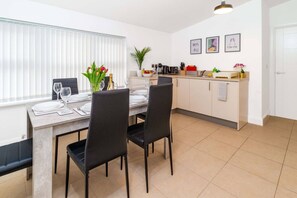 A dining area with a table set for four, a kitchen with white cabinets, and a window with blinds.