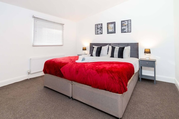 A bedroom with a bed, bedside tables, a radiator, a window with blinds, and wall art.