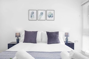 A bedroom with a bed, two nightstands, two lamps, a window with blinds, and three framed leaf artworks on the wall.