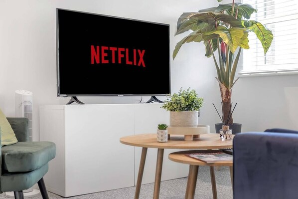 Un salon moderne avec un téléviseur à écran plat fixé au mur, affichant le logo « NETFLIX », un fauteuil vert, une table ronde en bois sur laquelle est posée une plante en pot, et une grande plante en pot dans un coin.
