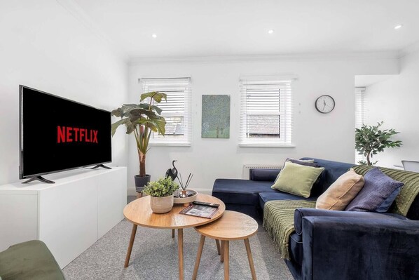 Un salon moderne avec un téléviseur à écran plat affichant le logo « NETFLIX », un canapé bleu foncé, une table basse ronde en bois et deux plantes en pot.