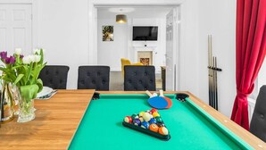 A pool table with billiard balls, a vase of flowers, and a dining table with chairs in a room with white walls and a red curtain. - The No.7 | Modern 3BR House | Pool Table Pingpong (Brampton)
