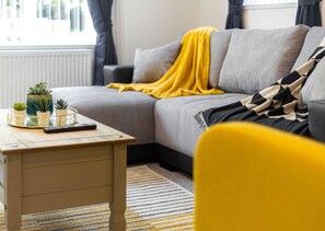 A living room with a sofa, a yellow chair, a coffee table with plants and a remote control, and a striped rug.