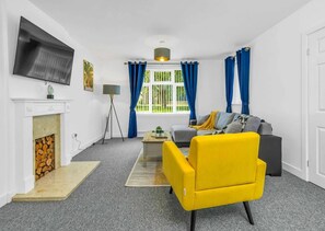 Una sala de estar con chimenea, un sofá, un sillón amarillo y un televisor montado en la pared.