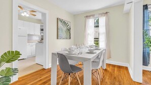 Comedor con una mesa dispuesta para seis personas, armarios blancos, ventilador de techo y ventana con cortinas livianas.