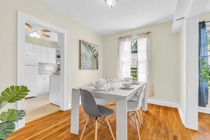 Comedor con una mesa dispuesta para seis personas, armarios blancos, ventilador de techo y ventana con cortinas livianas.