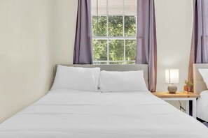 A neatly made bed with white linens, a bedside table with a lamp and a small plant, and a window with curtains. - Cute Bungalow near Downtown, Sleeps 6 (Spartanburg)