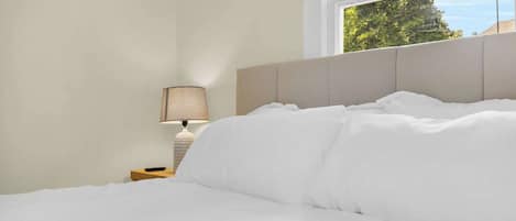A neatly made bed with white bedding, a bedside lamp, and a window with a view of trees.