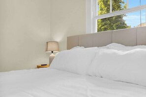 A neatly made bed with white bedding, a bedside lamp, and a window with a view of trees.