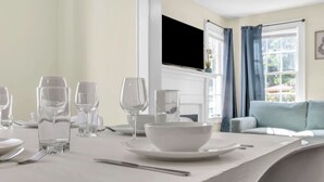 A dining table set with clear glassware and white plates, a white bowl, and a spoon, with a window and curtains in the background.