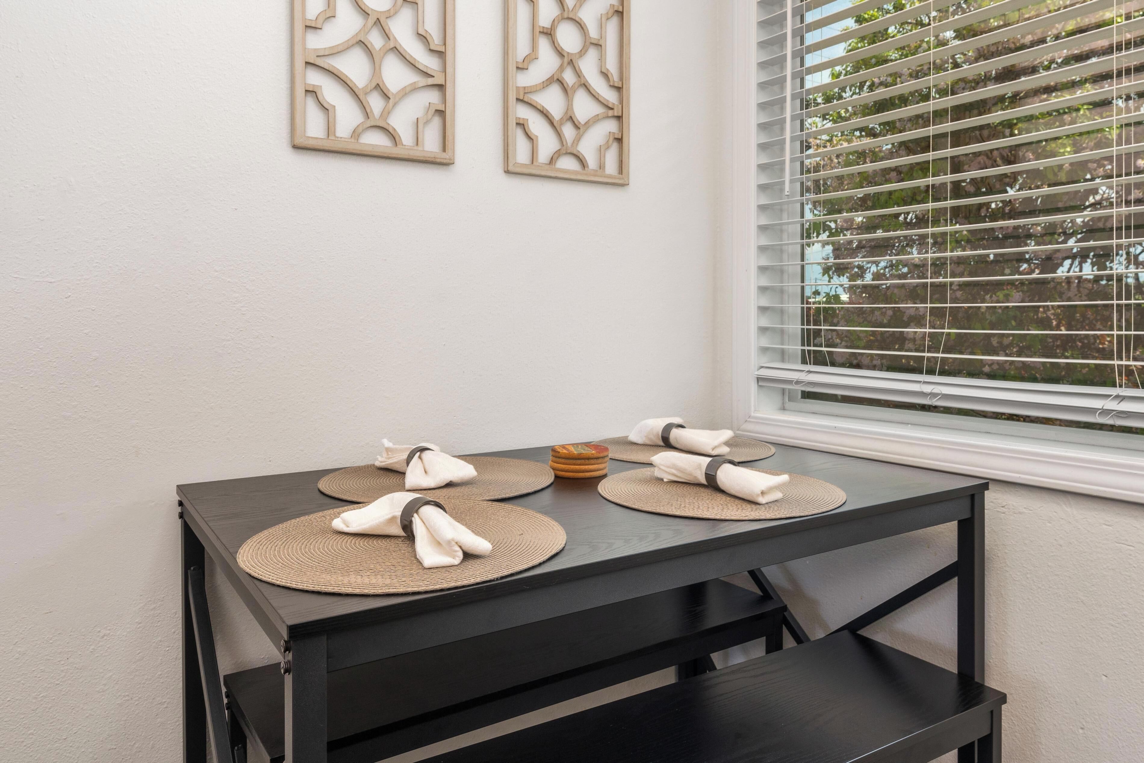 Een eettafel met placematen en opgevouwen servetten, voor een witte muur met decoratieve panelen en een raam met zonwering.