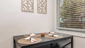 Una mesa de comedor con individuales y servilletas dobladas, frente a una pared blanca con paneles decorativos y una ventana con persianas.