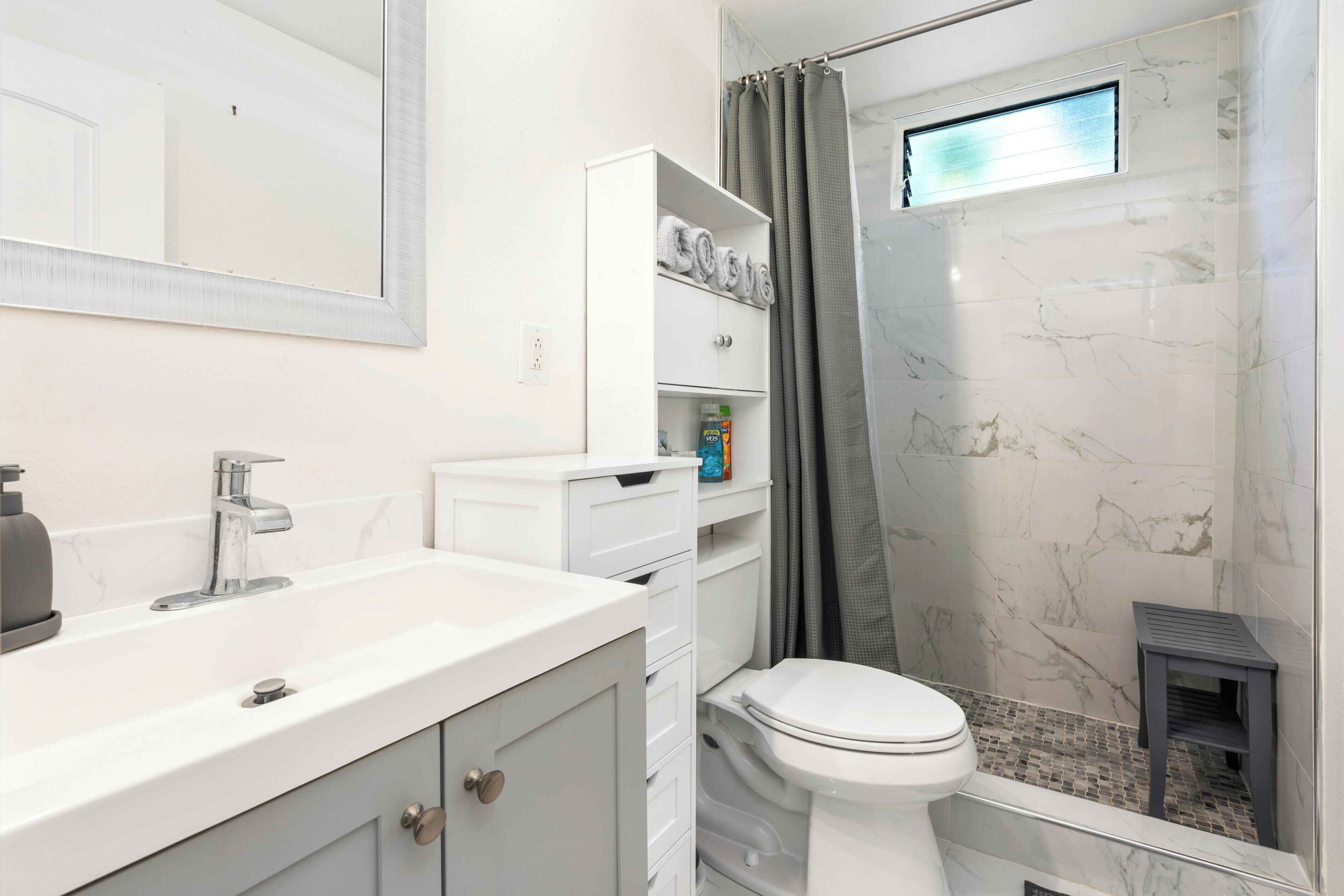 Un baño con lavamanos blanco, inodoro, ducha con puerta de cristal y un pequeño estante con artículos de aseo.