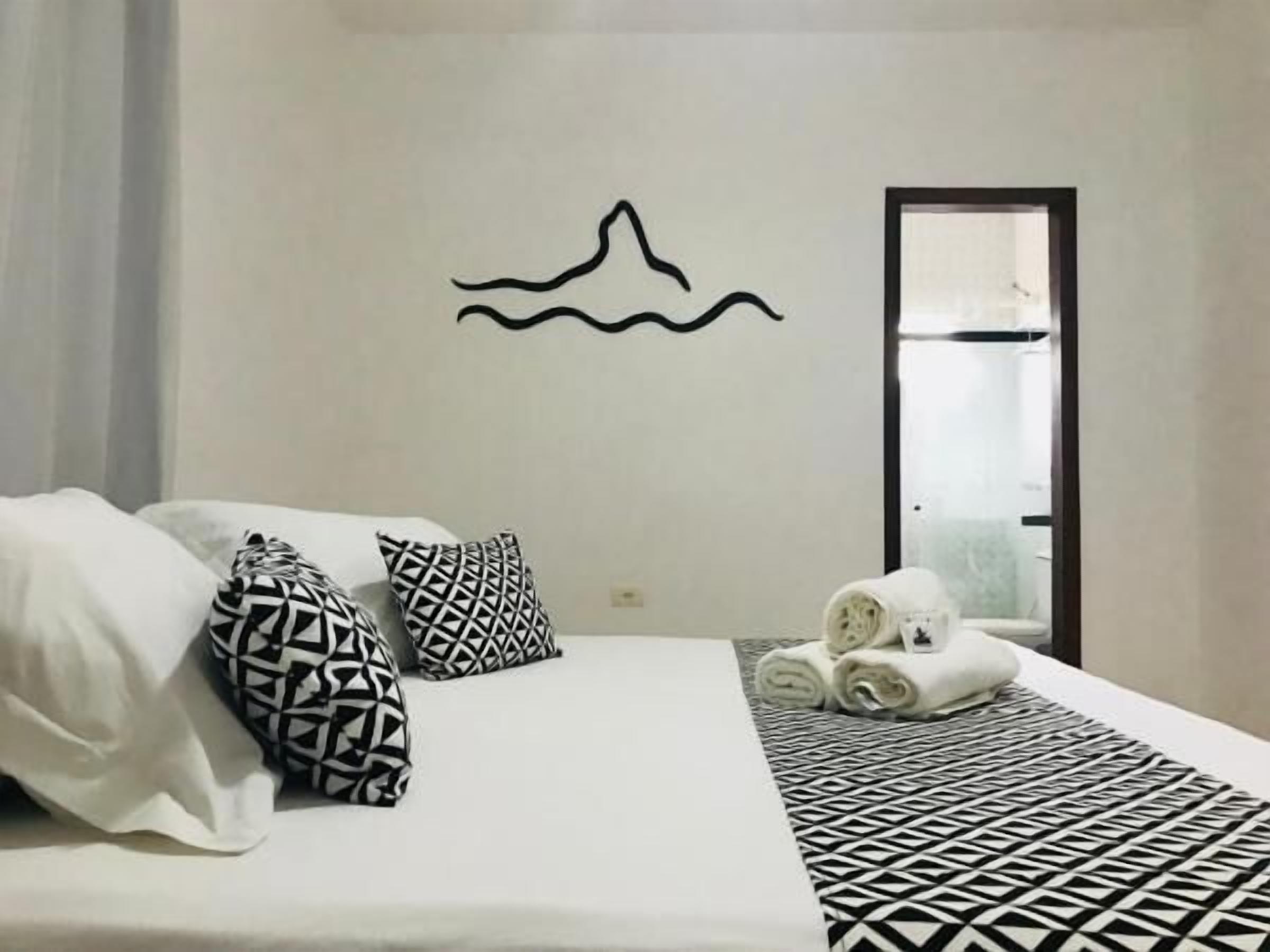 Habitación de hotel con cama, almohadas estampadas, un perchero con toallas y un adorno de pared.