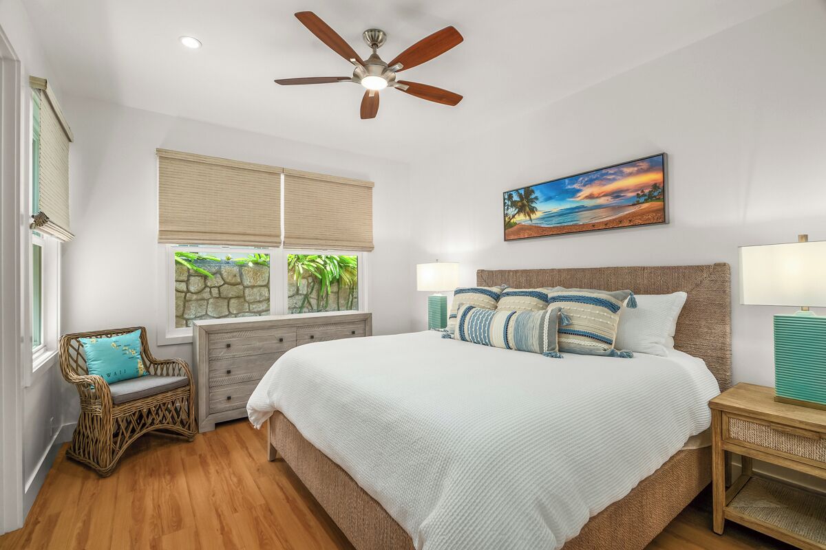 A bedroom with a large bed, a ceiling fan, a window with blinds, a wicker chair, a nightstand, and a painting on the wall.