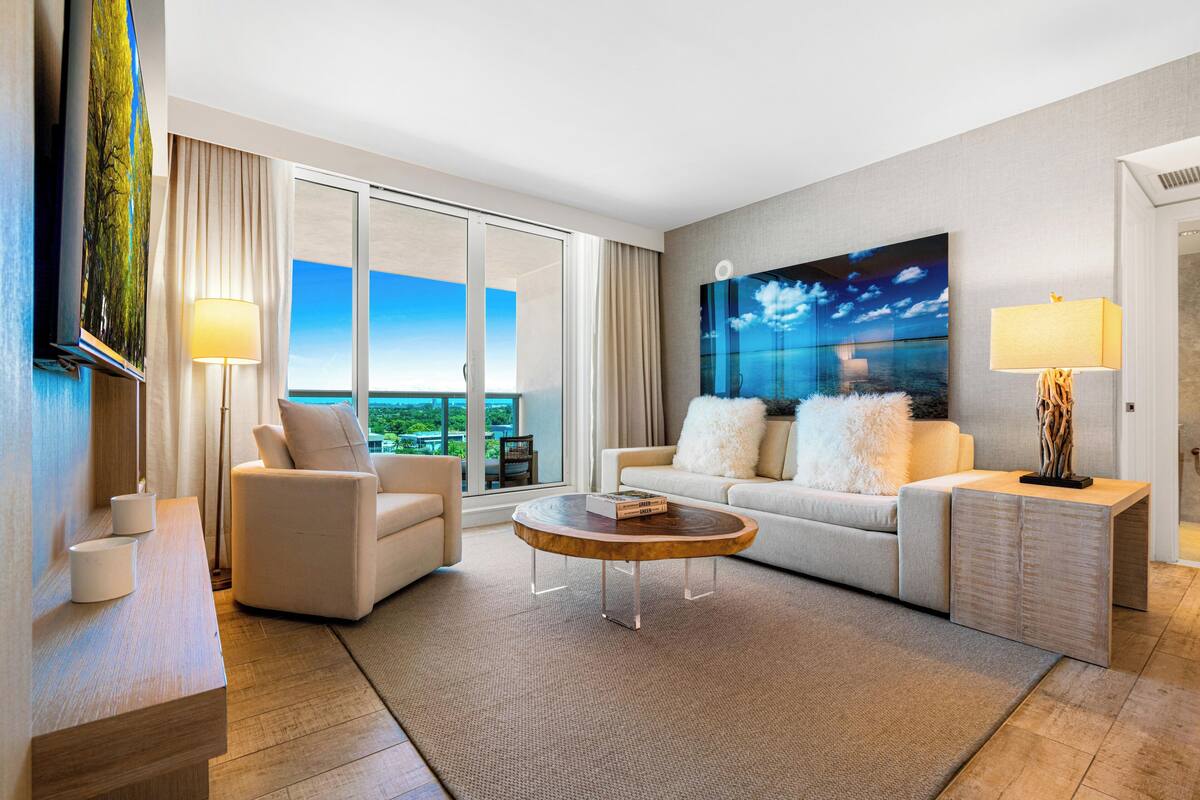 A modern hotel room with a large window, a sofa, a coffee table, and a TV.