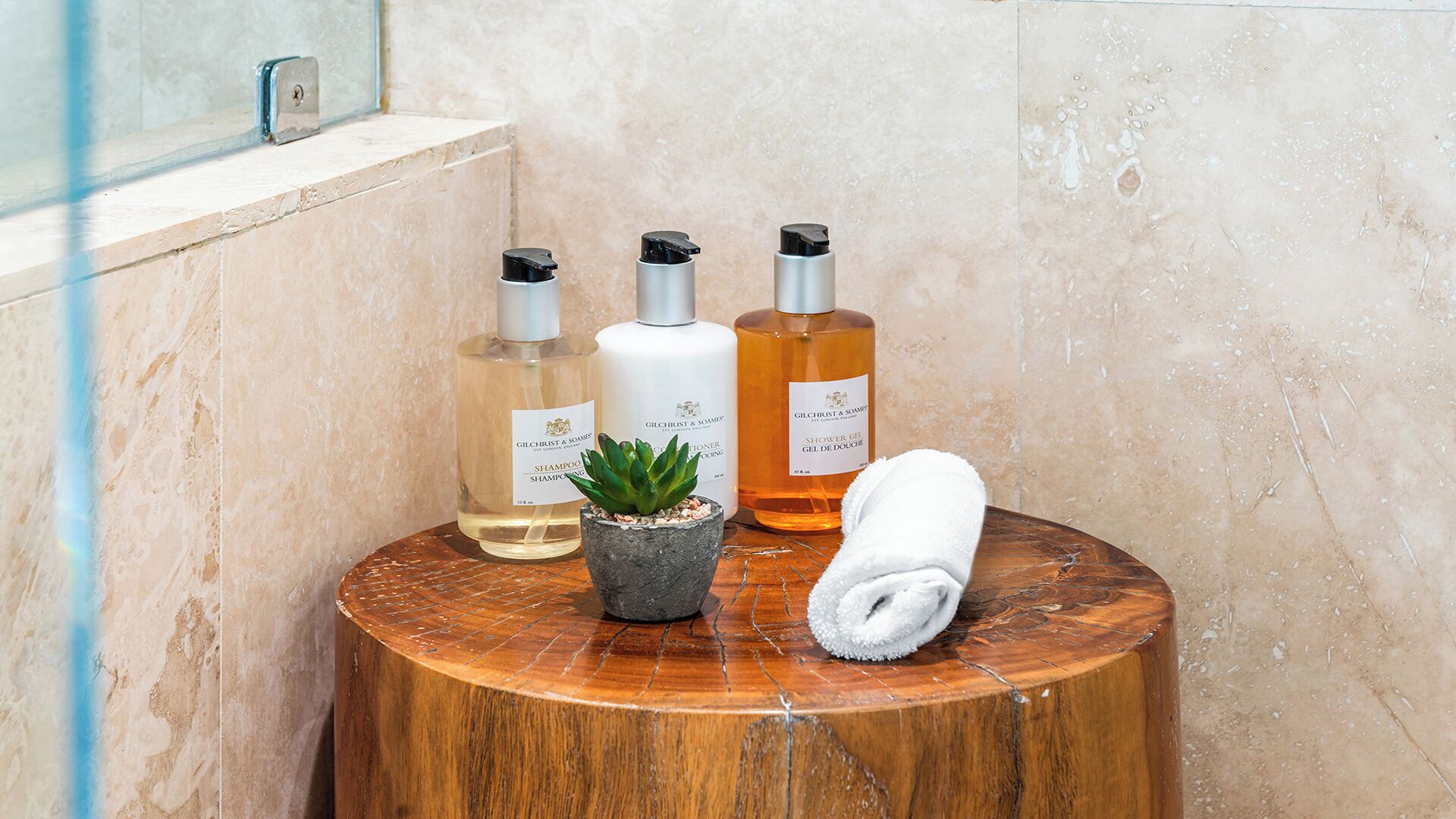 Un tocador de baño con tres botellas de productos Gilchrist & Somers, una pequeña planta en maceta y una toalla enrollada sobre un taburete de madera.