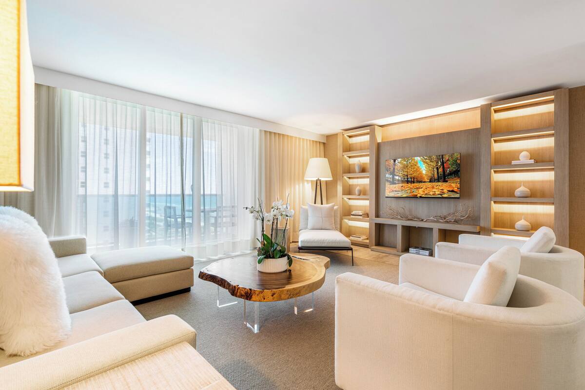 A modern living room with a large window, a wooden coffee table, white sofas, a TV, and a bookshelf.