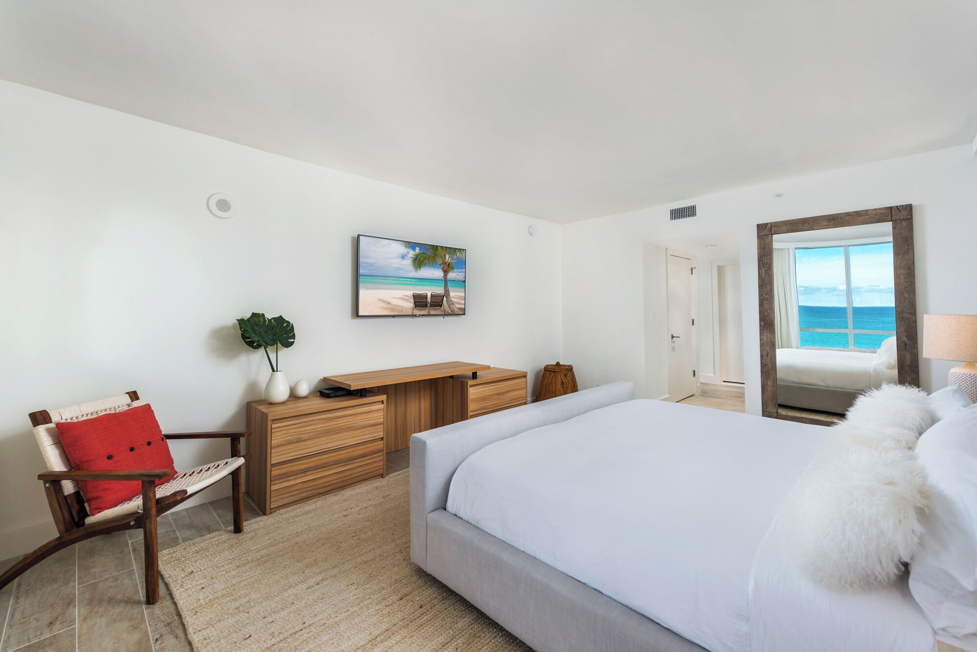 A bedroom with a bed, a desk, a chair, a large window with a view of the sea, and a painting on the wall.