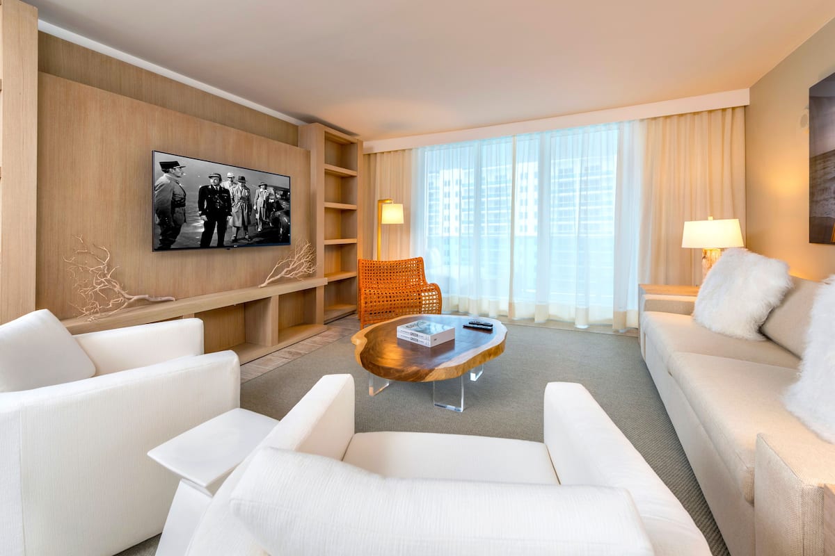 A modern hotel room with a white sofa, a wooden coffee table, a television mounted on the wall, and a large window with sheer curtains.